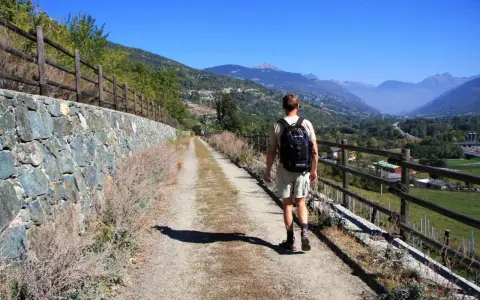 Man vandrar i Italien på en grusväg med stenmur och staket. Vacker bergig dal i bakgrunden.