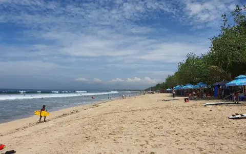 En man med en gul surfbräda går längs Kuta Beach. Vågor rullar in mot stranden där folk solar och surfar.