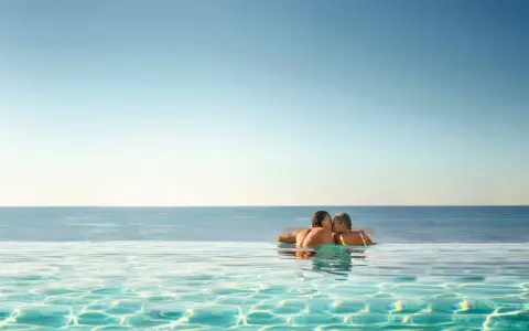 Ett par kysser varandra i en infinitypool med havet i bakgrunden. Perfekt för romantiska resmål.