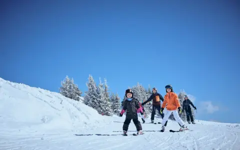 Familj njuter av skidåkning i mars. Barn och vuxna i skidkläder på en snöig backe med träd i bakgrunden. Ett perfekt resmål mars för vinteraktiviteter.