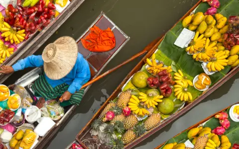 En säljare i en båt på en flytande marknad i Thailand, omgiven av ett överflöd av färsk frukt som bananer, mango och ananas.