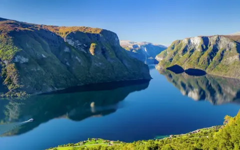 En båt färdas på en spegelblank fjord i Norge, omgiven av gröna bergssluttningar. En tågresa genom detta landskap vore en dröm.
