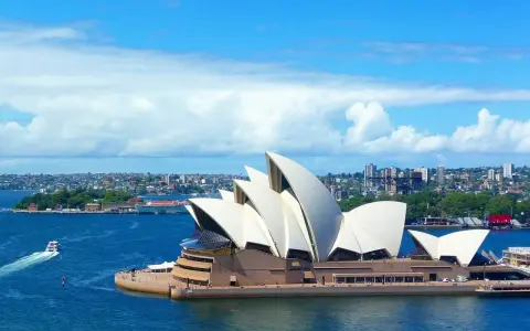Sydney stad med operahuset i förgrunden och en båt på det blå vattnet.