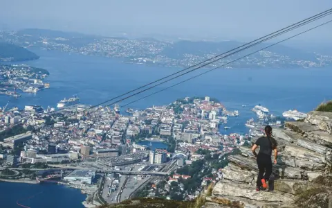 En kvinna står på en klippa och blickar ut över en stad vid havet, med berg i bakgrunden.