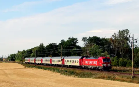 Ett rött lok drar ett långt tåg med vita vagnar genom ett landskap med guldigt höstfält och gröna träd. Kanske är det ett nattåg till Berlin.