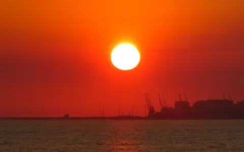 En glödande sol går ner över havet, med silhuetter av hamnkranar och byggnader i fjärran. En av de fina solnedgångar som får en att drömma sig bort.