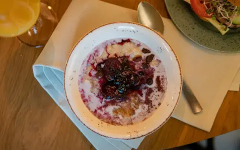 En skål med gröt toppad med bär och russin, en del av en kroatisk frukost. Bredvid syns en smörgås och ett glas juice.