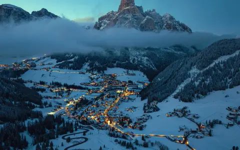En snötäckt by i Italien på vintern, upplyst av tusentals ljus. Majestätiska berg reser sig bakom, inbäddade i dimma.