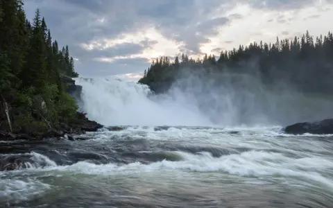 Vackra vattenfall dånar nerför klipporna, omgivna av en tät grön skog under en dramatisk molnhimmel.