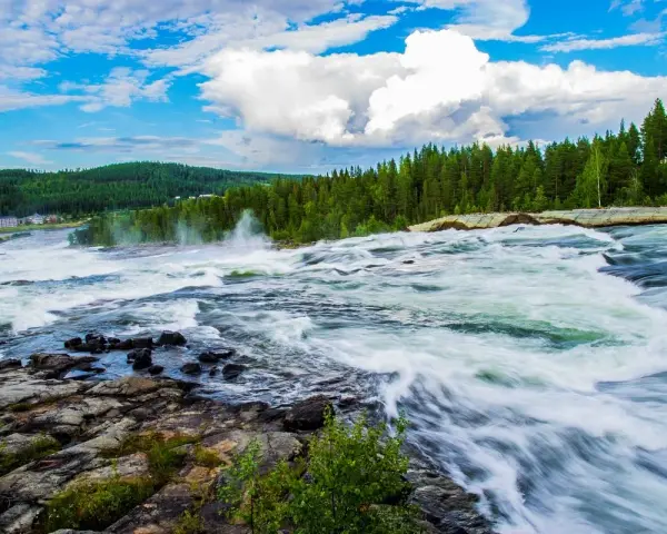 Vattenfall i Luleå med forsande vatten, grönskande skog och en blå himmel med vita moln.
