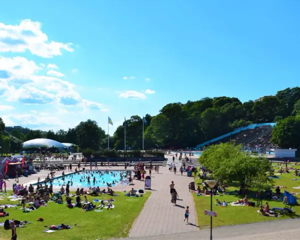 En solig dag vid en populär badplats i Stockholm. Människor solar, badar och njuter av sommaren på gräsmattor och i poolen.