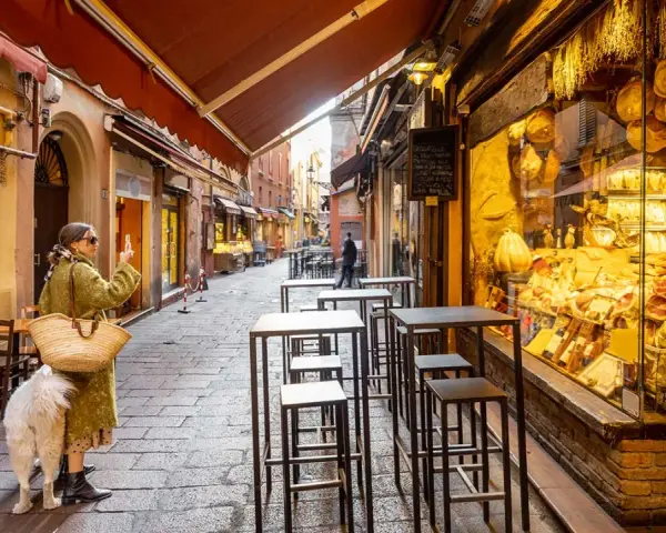 En kvinna med hund tar bild på en charmig gränd i Bologna, Italien, med ett skyltfönster fyllt av läcker bologna mat.