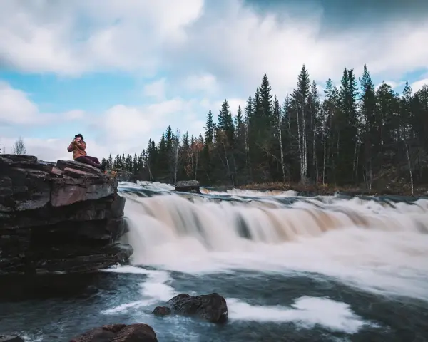 Person sitter på en klippa vid ett forsande vattenfall i Dalarna, ett perfekt utflyktsmål.