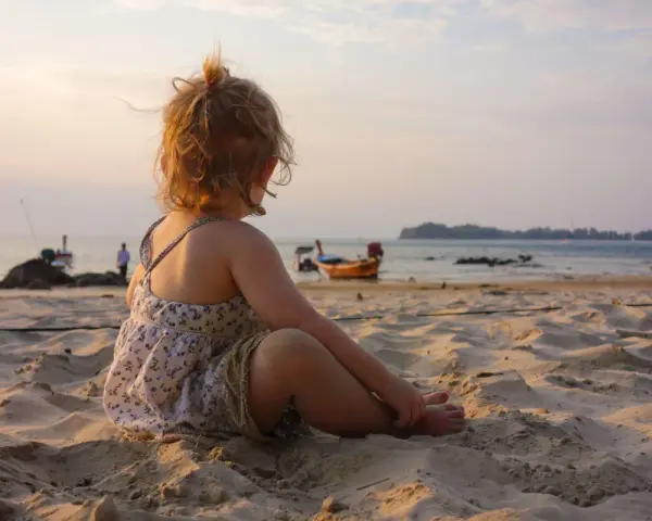 Liten flicka sitter på stranden och tittar ut över havet. Det är varmt på vintern, med båtar i bakgrunden och en vacker solnedgång.