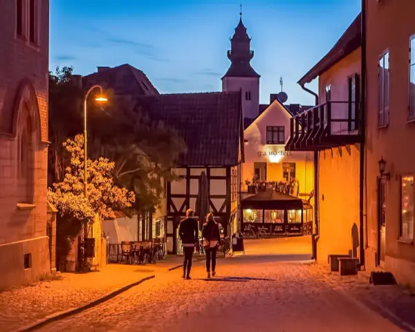 Två personer promenerar på en kullerstensgata i Båstad under Stockholmsveckan. Kvällen är ljum, och husen längs gatan lyser varmt.