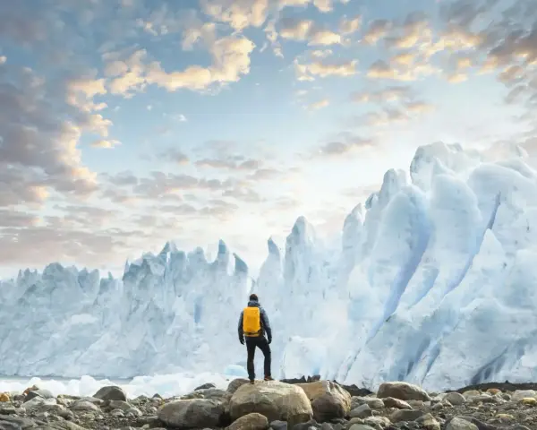 En person med gul ryggsäck står på stenar framför en enorm glaciär. Ett perfekt resmål i januari för den äventyrlige.