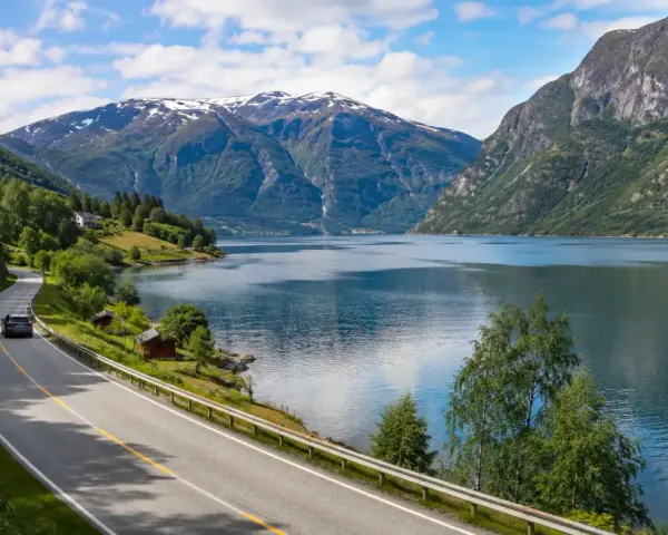 En bil kör längs en slingrande väg vid en fjord, med snöklädda berg i bakgrunden. En perfekt bild för en semester i Norge.