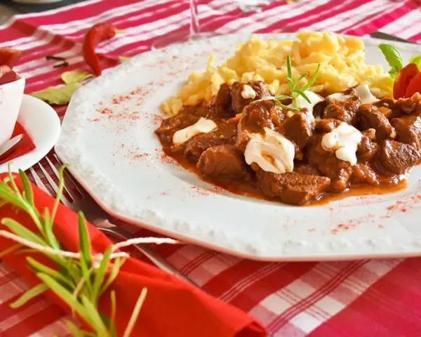 En smak av mat i Budapest: mustig gulasch med nokedli, garnerad med gräddfil och en rosmarinkvist. En röd chili och en tomatblomma fulländar upplägget.