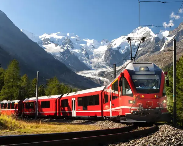 Ett rött tåg slingrar sig fram genom ett dramatiskt alplandskap med snöklädda berg. Kanske är det ett tåg till Italien, med hisnande vyer längs vägen.