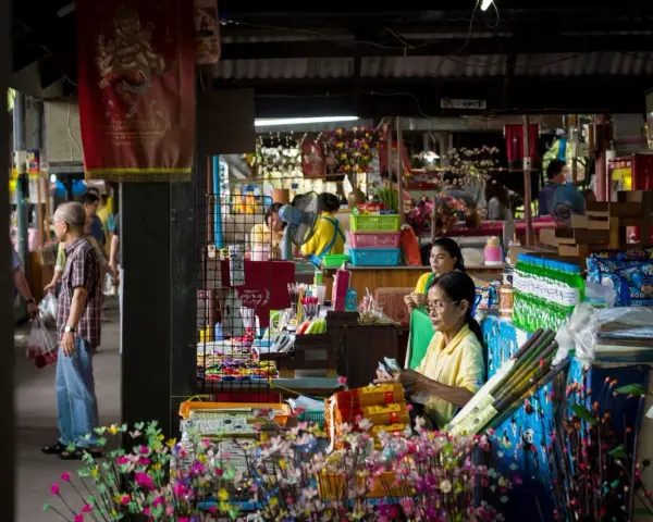 En livlig marknad i Bangkok med försäljare och kunder bland färgglada varor och blommor.