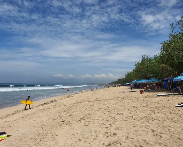 En man med en gul surfbräda går längs Kuta Beach. Vågor rullar in mot stranden där folk solar och surfar.