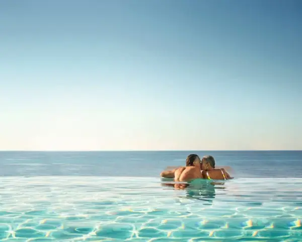 Ett par kysser varandra i en infinitypool med havet i bakgrunden. Perfekt för romantiska resmål.