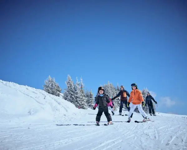 Familj njuter av skidåkning i mars. Barn och vuxna i skidkläder på en snöig backe med träd i bakgrunden. Ett perfekt resmål mars för vinteraktiviteter.