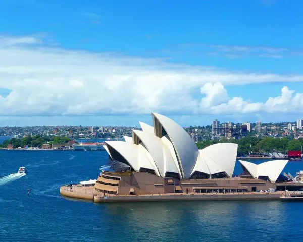 Sydney stad med operahuset i förgrunden och en båt på det blå vattnet.