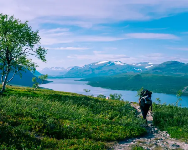 Vandrare med stor ryggsäck på en stenstig, njuter av storslagna vyer över sjö och snöklädda berg. Perfekt för vandringsleder Norrland.