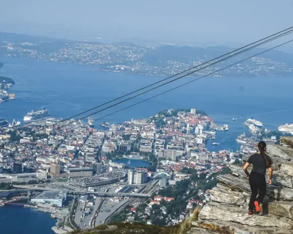 En kvinna står på en klippa och blickar ut över en stad vid havet, med berg i bakgrunden.