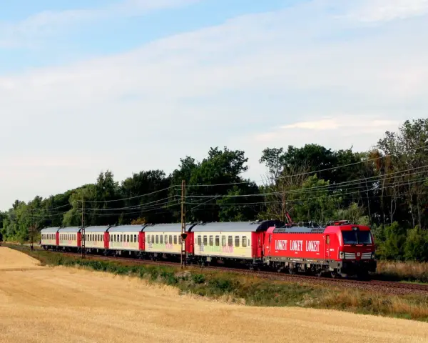 Ett rött lok drar ett långt tåg med vita vagnar genom ett landskap med guldigt höstfält och gröna träd. Kanske är det ett nattåg till Berlin.