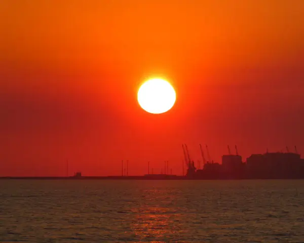 En glödande sol går ner över havet, med silhuetter av hamnkranar och byggnader i fjärran. En av de fina solnedgångar som får en att drömma sig bort.