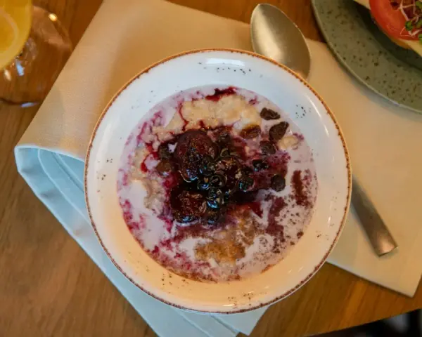 En skål med gröt toppad med bär och russin, en del av en kroatisk frukost. Bredvid syns en smörgås och ett glas juice.