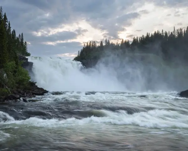 Vackra vattenfall dånar nerför klipporna, omgivna av en tät grön skog under en dramatisk molnhimmel.