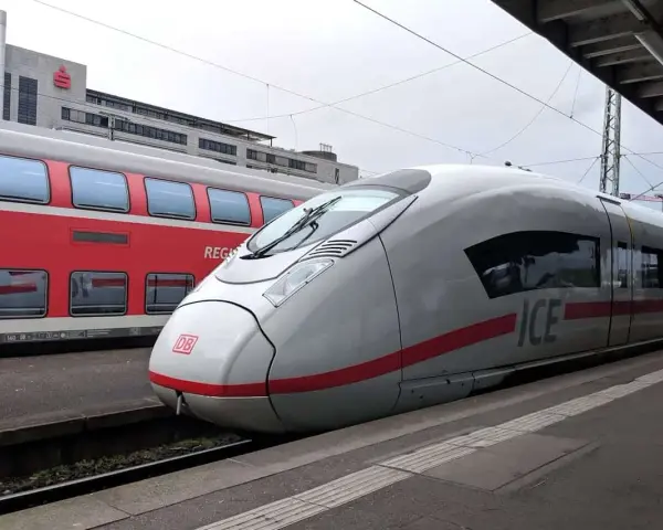 Ett modernt ICE-tåg står redo vid perrongen, perfekt för en resa med tåg till Tyskland. Bredvid syns ett rött dubbeldäckartåg.