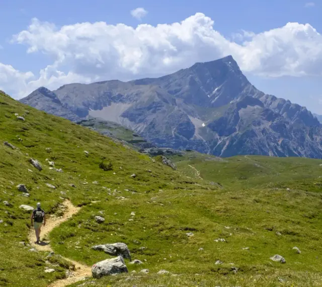Vandrare på en stig i Dolomiterna, omgiven av gröna sluttningar och majestätiska berg under en molnig himmel.