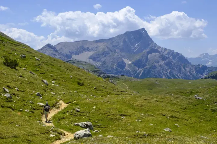 Vandrare på en stig i Dolomiterna, omgiven av gröna sluttningar och majestätiska berg under en molnig himmel.