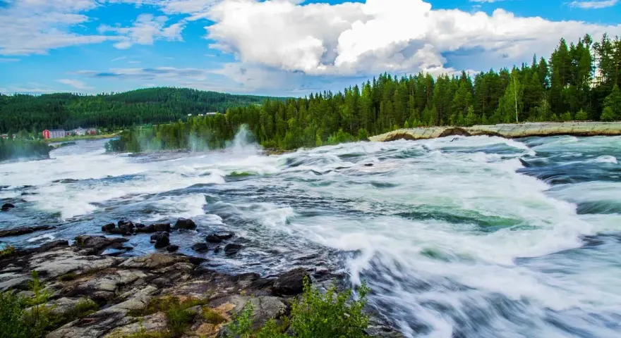 Vattenfall i Luleå med forsande vatten, grönskande skog och en blå himmel med vita moln.