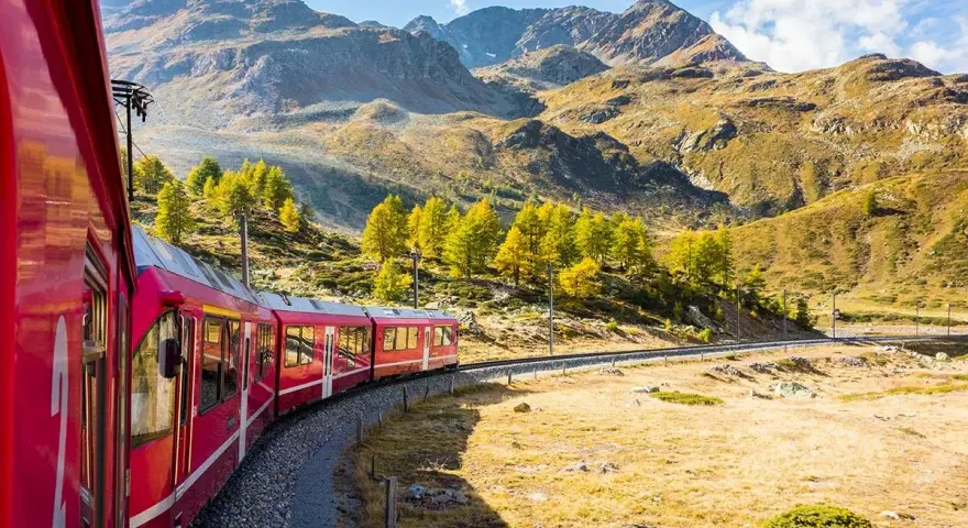 Ett rött tåg slingrar sig genom ett höstfärgat landskap med berg i bakgrunden. Perfekt för att tågluffa i Sverige och uppleva naturens skönhet.
