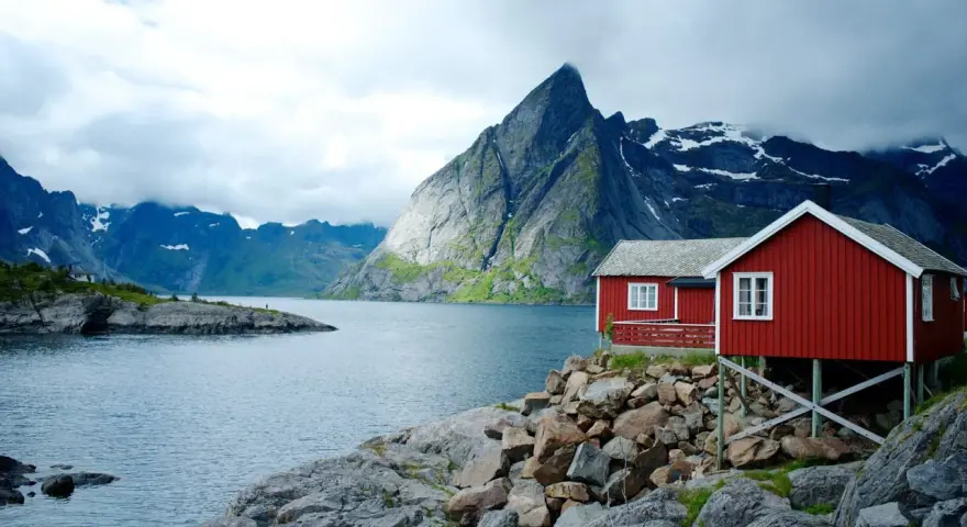 Två röda stugor vid en sjö i Norge, med dramatiska berg i bakgrunden. Perfekt för den som vill vandra i Norge.