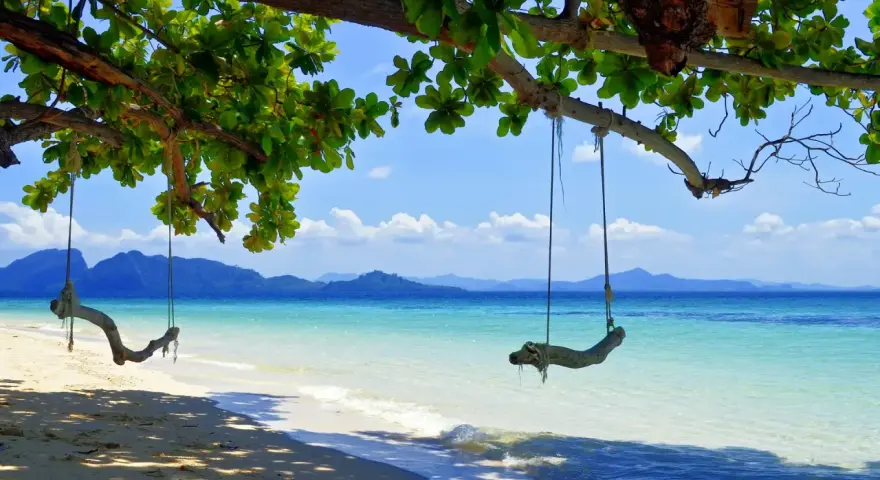 Gungor hänger från ett träd vid en strand i Thailand. Vackert turkost vatten och berg i bakgrunden. En idyllisk plats, trots att det är regnperiod.
