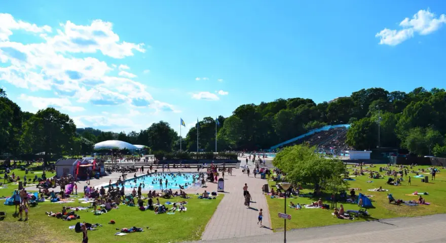En solig dag vid en populär badplats i Stockholm. Människor solar, badar och njuter av sommaren på gräsmattor och i poolen.