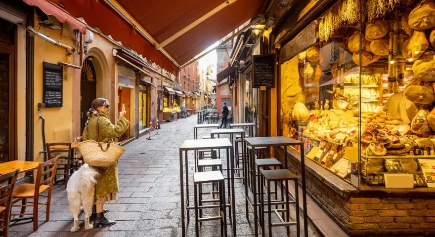 En kvinna med hund tar bild på en charmig gränd i Bologna, Italien, med ett skyltfönster fyllt av läcker bologna mat.