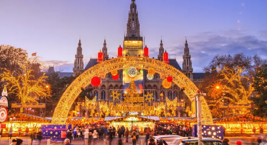 En magisk julmarknad i Wien, med Rådhuset i bakgrunden, gnistrar av tusentals ljus. En båge av gyllene ljusslingor välkomnar besökare till den livliga julmarknaden.