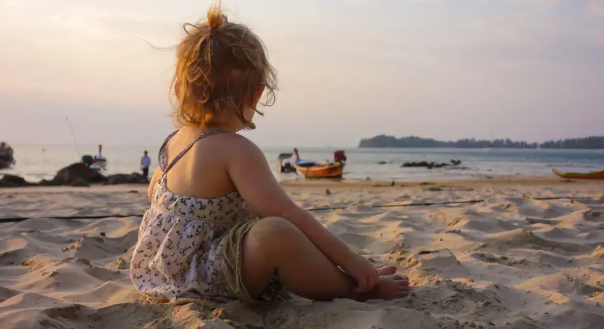 Liten flicka sitter på stranden och tittar ut över havet. Det är varmt på vintern, med båtar i bakgrunden och en vacker solnedgång.