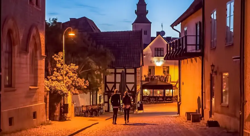 Två personer promenerar på en kullerstensgata i Båstad under Stockholmsveckan. Kvällen är ljum, och husen längs gatan lyser varmt.