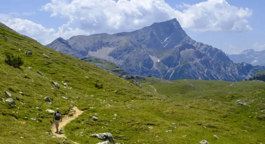 Vandrare på en stig i Dolomiterna, omgiven av gröna sluttningar och majestätiska berg under en molnig himmel.