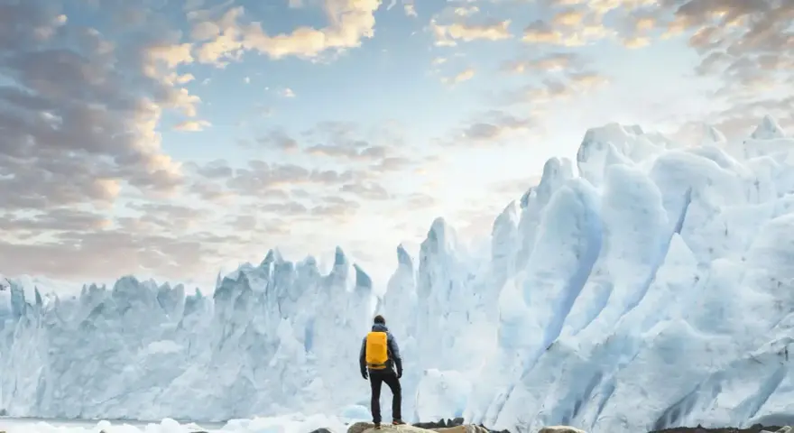 En person med gul ryggsäck står på stenar framför en enorm glaciär. Ett perfekt resmål i januari för den äventyrlige.