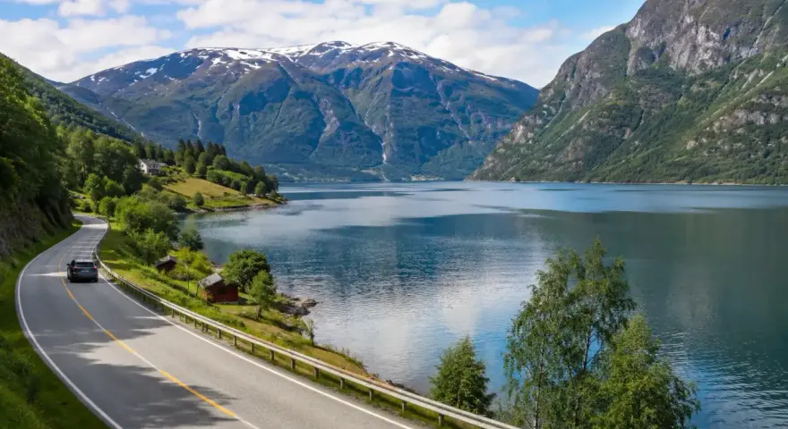 En bil kör längs en slingrande väg vid en fjord, med snöklädda berg i bakgrunden. En perfekt bild för en semester i Norge.