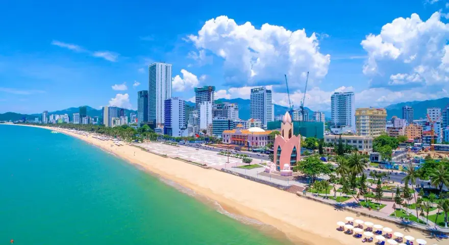 En vidsträckt strand med turkost vatten och en stadssilhuett i bakgrunden. Nha Trang beach är en populär destination.